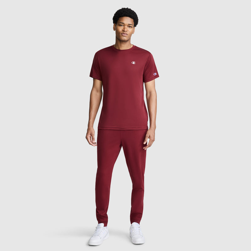 A man with short, curly hair faces forward wearing a matching maroon Champion Sport T-Shirt, Reflective C Logo, and joggers. He pairs the outfit with white sneakers. The men’s athletic shirt from Champion features moisture-wicking technology to keep him comfortable and dry. The background is plain and light gray.