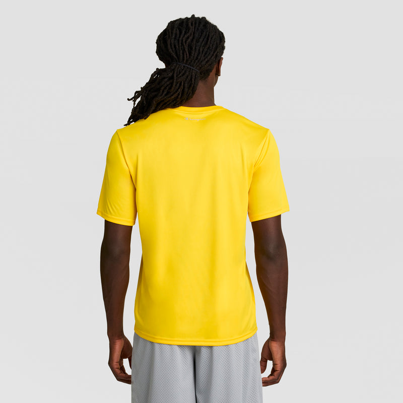 A person with long, dark dreadlocks stands facing away, wearing a bright yellow Champion Sport T-Shirt with a Reflective C Logo and light gray shorts against a plain light gray background.