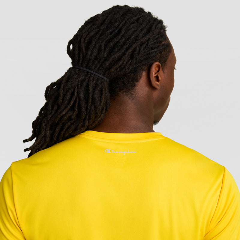 A person with long, dark dreadlocks tied back is seen from behind wearing a bright yellow Champion Sport T-Shirt with a Reflective C Logo, against a plain light gray background.