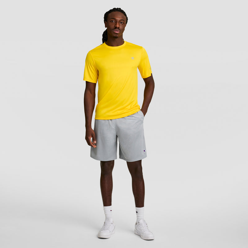 A man faces forward against a light gray background, wearing a bright yellow Champion Sport T-Shirt with a reflective C logo and Double Dry tech, paired with light gray shorts, white crew socks, and white sneakers. His right hand is in his pocket.