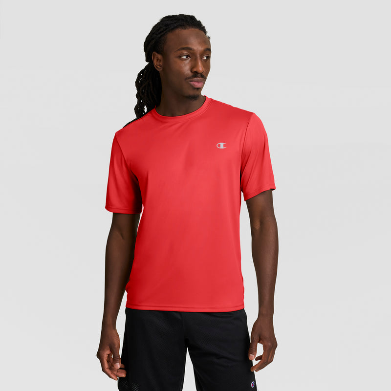 A man with long braided hair wears a bright red Champion Sport T-Shirt with a reflective C logo and black shorts, standing against a light gray background while looking to his left.