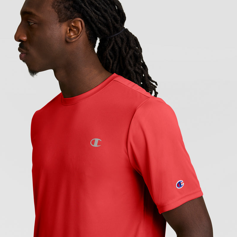 A man with long, dark dreadlocks is seen from the side wearing a bright red Champion Sport T-Shirt with Double Dry technology and a reflective C logo, set against a plain light gray background.