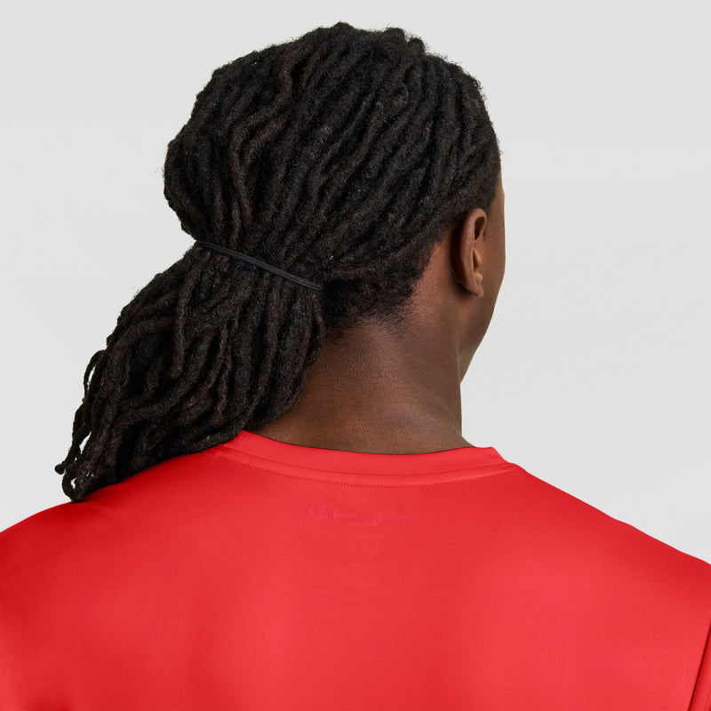 A person with long black dreadlocks tied back wears a bright red Champion Sport T-Shirt with Reflective C Logo, shown from behind against a plain light gray background.
