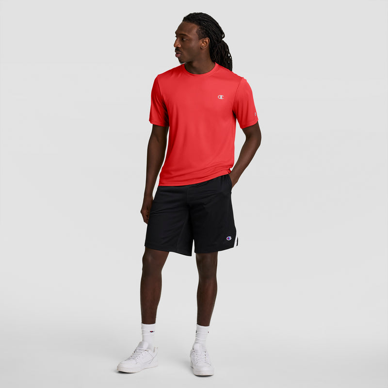 A man wears a bright red Champion Sport T-Shirt with Reflective C Logo, black shorts, white socks, and white sneakers, standing against a light gray background with one hand in his pocket.