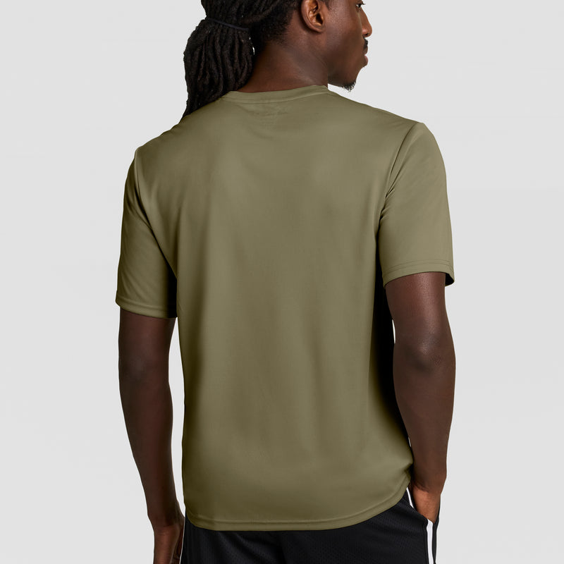 A man with long braided hair, seen from behind, wears a Champion Sport T-Shirt with Double Dry technology and a reflective C logo in olive green, paired with black shorts, standing against a plain light gray background.