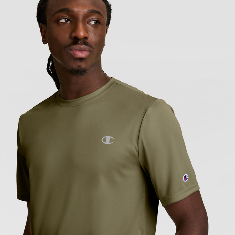 A man with long braided hair wears an olive green Champion Sport T-Shirt with a reflective C logo on the chest and sleeve, featuring Double Dry technology. He looks to the side against a plain, light background.