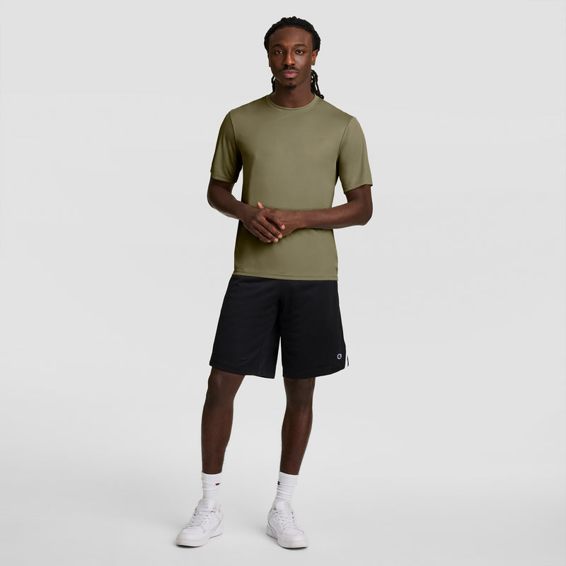 A man faces the camera with hands clasped, wearing a Champion Sport T-Shirt with Reflective C Logo and Double Dry tech in olive green, black athletic shorts, white socks, and white sneakers. He has long braided hair and stands against a plain background.
