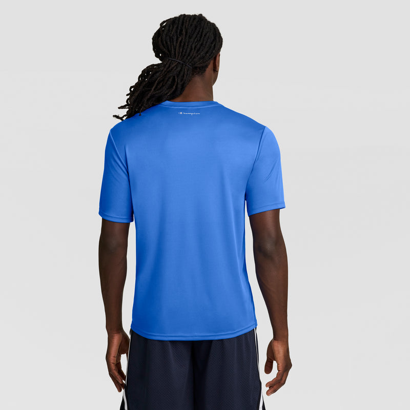 A man with long, dark braided hair is wearing a bright blue Champion Sport T-Shirt with Reflective C Logo and black shorts, standing facing away against a light gray background and displaying a modern athletic look.