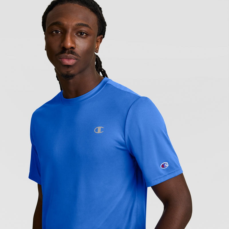 A man with long braided hair wears a bright blue Champion Sport T-Shirt featuring a reflective C logo, standing against a plain light gray background and looking at the camera.