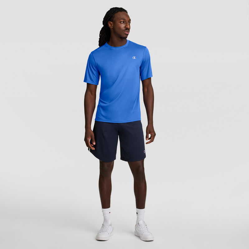 A man stands against a light gray background in an athletic look, wearing a blue Champion Sport T-Shirt with Reflective C Logo, navy shorts, white socks, and white sneakers. He looks to the side with arms relaxed at his sides.