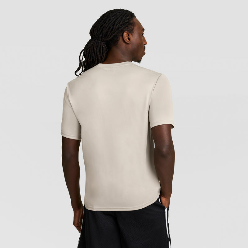 A person with long, braided hair, wearing a Champion Sport T-Shirt with Reflective C Logo and black shorts, stands facing away with hands in pockets against a plain light gray background.
