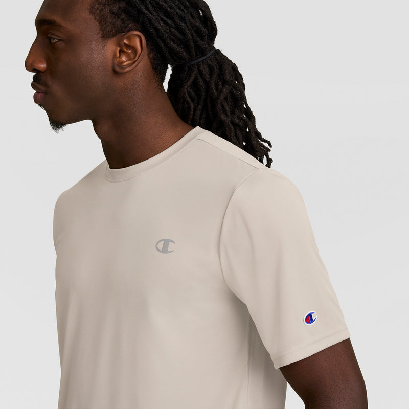 A man with long dark dreadlocks wears a light beige Champion Sport T-Shirt, Reflective C Logo. The logo appears on the chest and sleeve as he faces left against a plain light gray background.