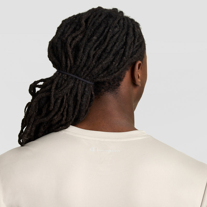 A person with long, dark dreadlocks tied back in a black hairband is seen from behind, wearing a light beige Champion Sport T-Shirt with a Reflective C Logo, against a plain light background.