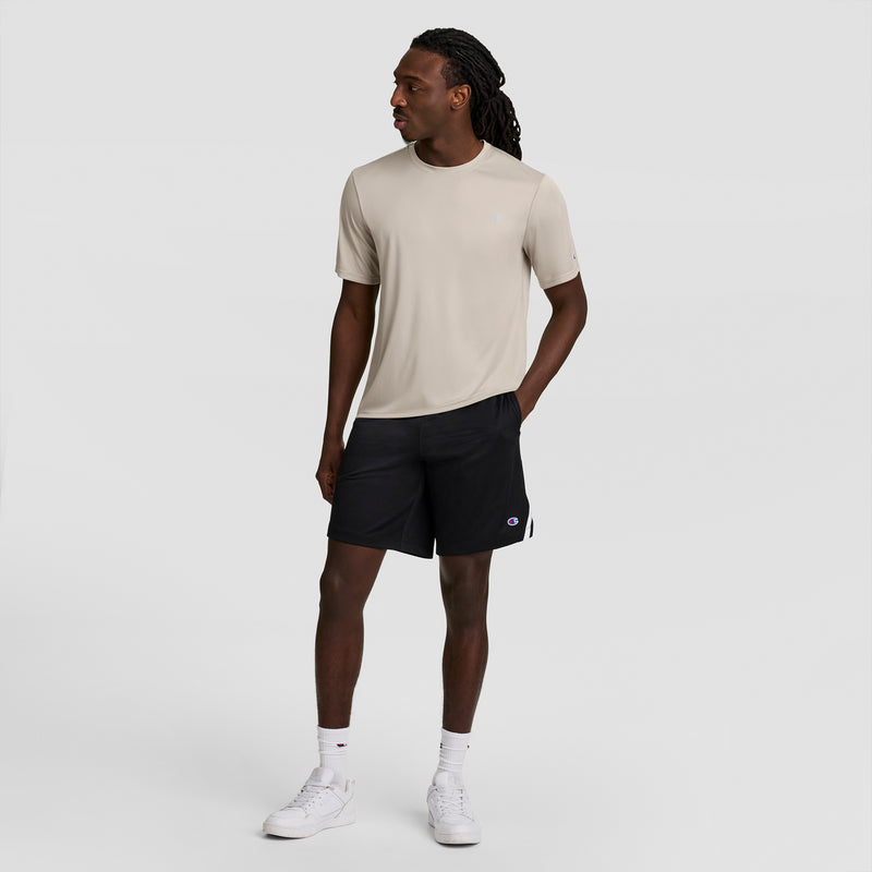 A man with long braided hair stands against a plain light backdrop, wearing a Champion Sport T-Shirt with Reflective C Logo in beige, black shorts, white crew socks, and white sneakers. He is looking left with one hand in his pocket.