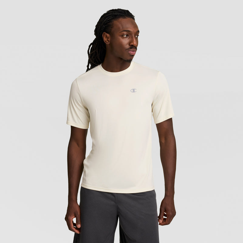 A man with long dreadlocks wears a cream Champion Sport T-Shirt featuring a reflective C logo and Double Dry tech, paired with dark shorts, standing against a plain light gray background.
