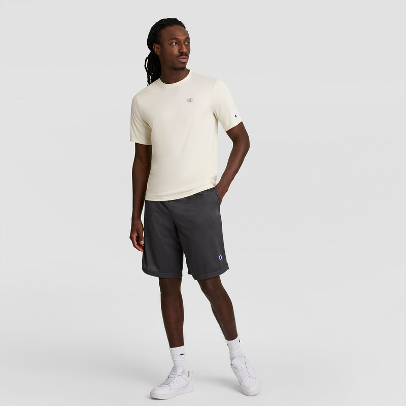 A man with long hair stands against a plain background, wearing a light beige Champion Sport T-Shirt with a reflective C logo and Double Dry tech, dark gray shorts, white crew socks, and white sneakers. His left hand is in his pocket.