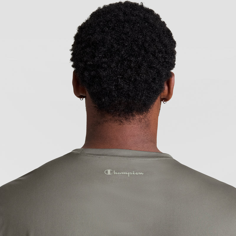 A person with short curly hair is shown from behind in a Champion Sport T-Shirt, Reflective C Logo - Big & Tall, olive green, featuring the logo on the upper back. The background is plain and light-colored.