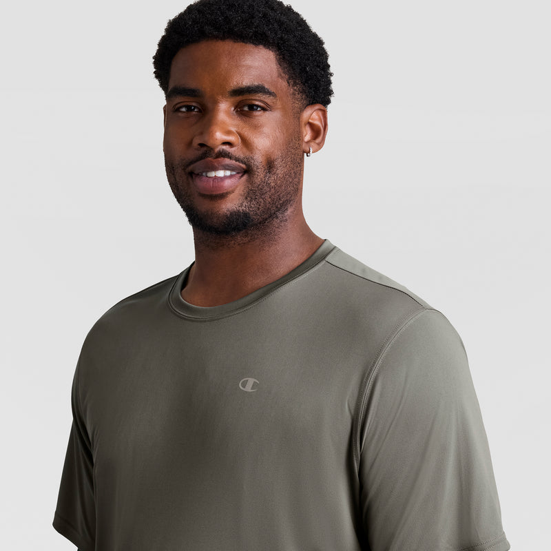 A man with short curly hair and a beard smiles while wearing a Champion Sport T-Shirt, Reflective C Logo - Big & Tall in olive green. The background is plain, light gray.