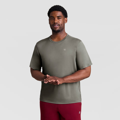 A man stands against a light gray background, wearing a Champion Sport T-Shirt, Reflective C Logo - Big & Tall in olive green with maroon shorts. He faces forward, smiling, with his hands loosely clasped.