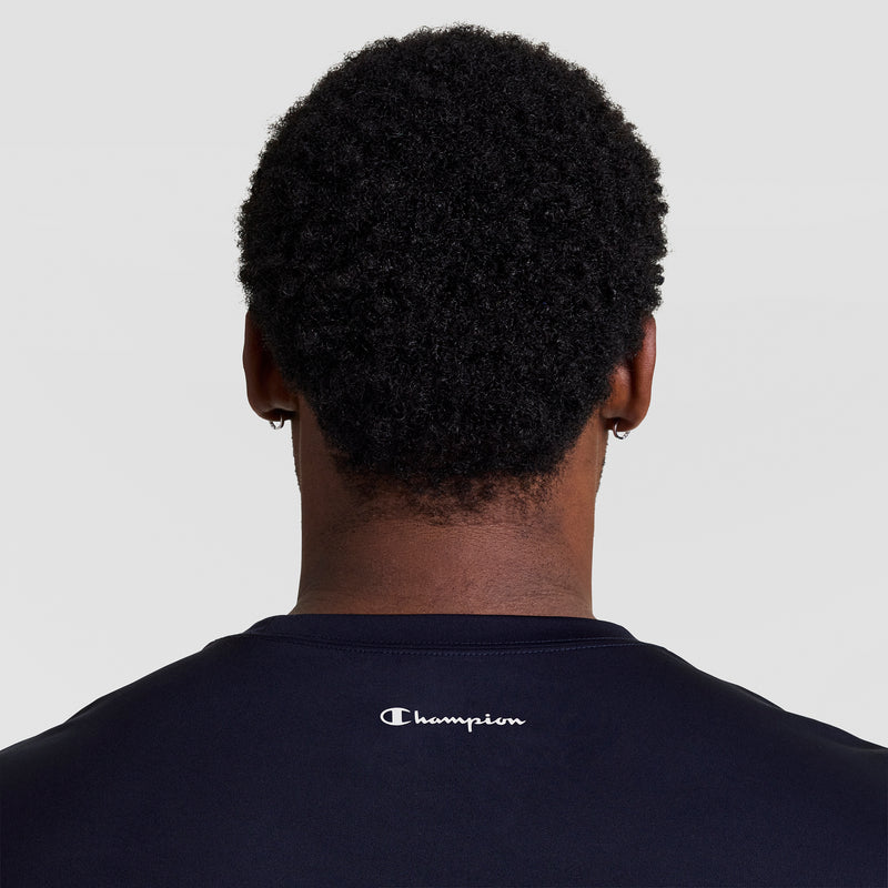 A person with short curly hair is seen from behind wearing a black Champion Sport T-Shirt, Reflective C Logo - Big & Tall, featuring the Champion name and logo in white on the upper back. The background is plain light gray.
