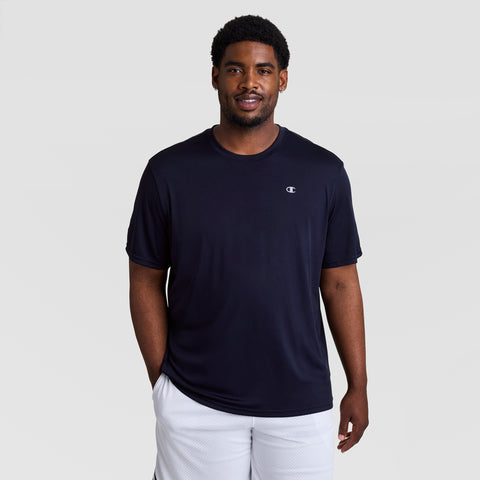 A man wears a Champion Sport T-Shirt, Reflective C Logo - Big & Tall in dark navy with a small logo on the chest and white athletic shorts, standing against a plain light gray background and smiling slightly.