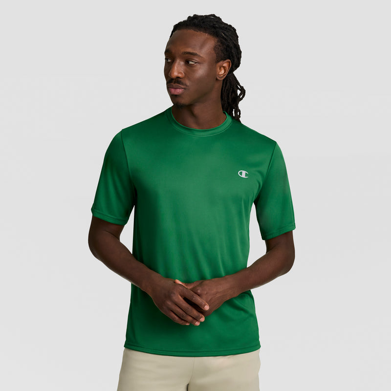 A man with long braided hair wears a green Champion Sport T-Shirt with a Reflective C Logo and Double Dry technology, paired with light-colored pants, standing against a plain light gray background with his hands loosely clasped.