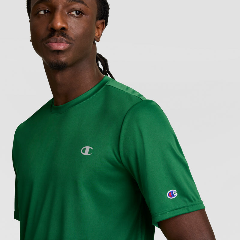 A man wears a green Champion Sport T-Shirt with reflective C logo on the chest and sleeve, standing against a plain light gray background.