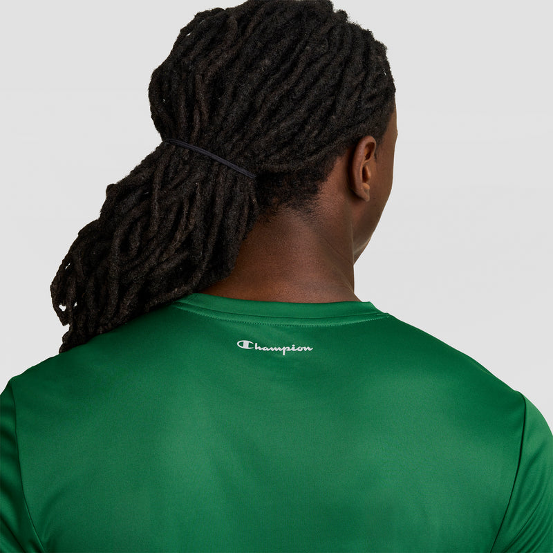 Seen from behind, a person with long dark dreadlocks wears a Champion Sport T-Shirt, Reflective C Logo—a green men’s short-sleeve athletic shirt with the white Champion emblem at the back—standing out against a plain light background.