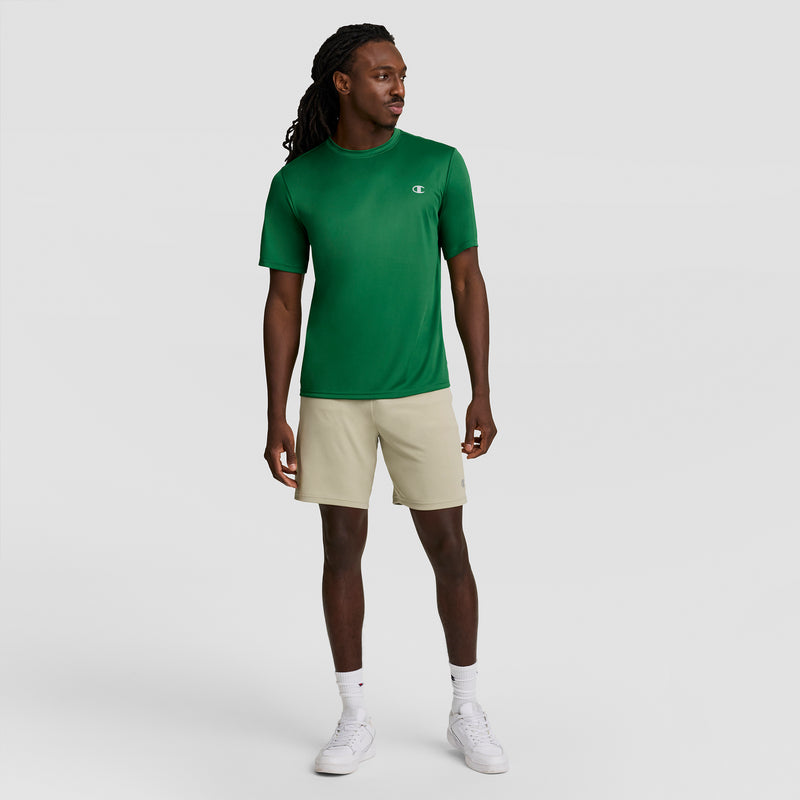 A man stands against a plain light gray background, wearing a green Champion Sport T-Shirt with Reflective C Logo and Double Dry technology, beige shorts, white crew socks, and white sneakers. He looks slightly right with arms relaxed at his sides.