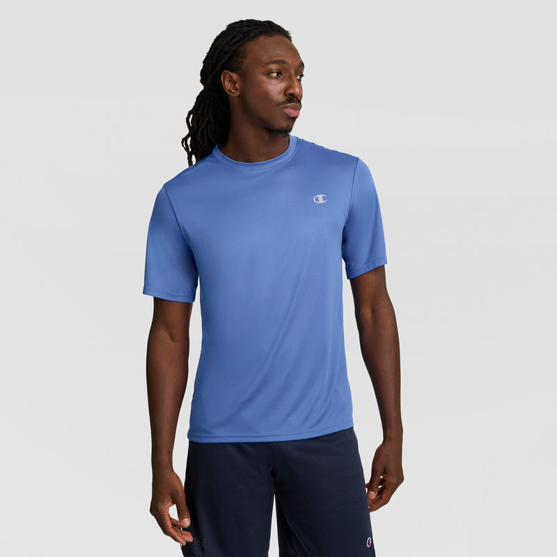 A man with long braided hair wears a Champion Sport T-Shirt in light blue featuring a Reflective C Logo and Double Dry technology, paired with navy shorts, as he stands against a plain gray background looking to his left.