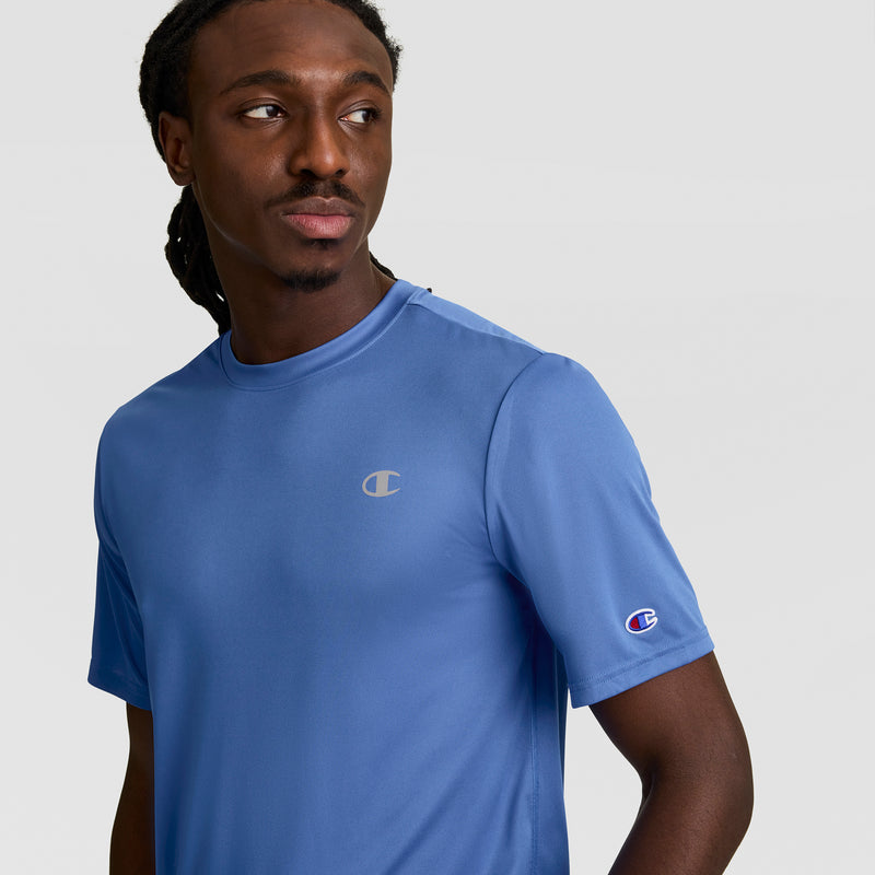 A man with long dark hair wears a Champion Sport T-Shirt, Reflective C Logo, in light blue. The short-sleeve tee features logos on the chest and sleeve and offers comfort with Double Dry technology. He looks left against a plain backdrop.