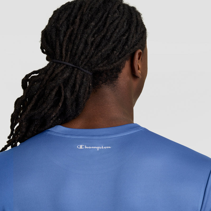 A person with long black dreadlocks tied back is seen from behind wearing a blue Champion Sport T-Shirt, Reflective C Logo. The white logo appears on the upper back against a plain, light-colored background.