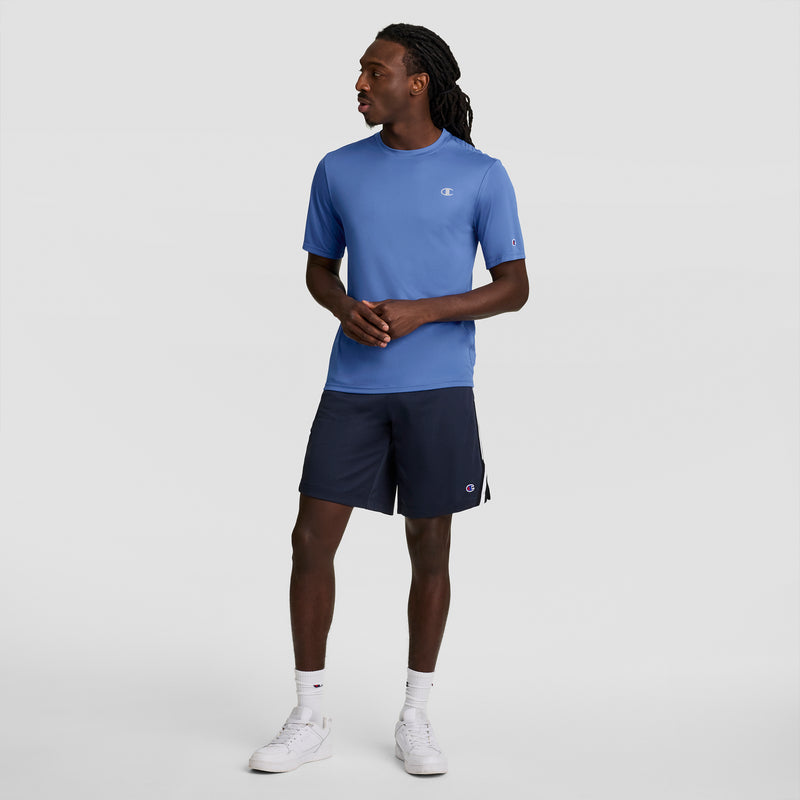 A man stands against a plain background wearing a Champion Sport T-Shirt with Reflective C Logo in light blue, dark navy shorts, white crew socks, and white sneakers. He looks to his left with his hands clasped.