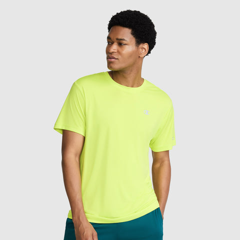 A man wearing a Champion Sport T-Shirt with a reflective C logo in bright yellow and teal shorts stands against a plain light gray background, looking slightly to his left with one hand in his pocket.