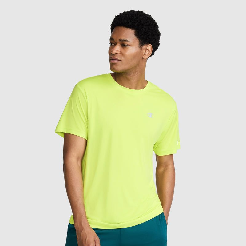 A man wearing a Champion Sport T-Shirt with a reflective C logo in bright yellow and teal shorts stands against a plain light gray background, looking slightly to his left with one hand in his pocket.
