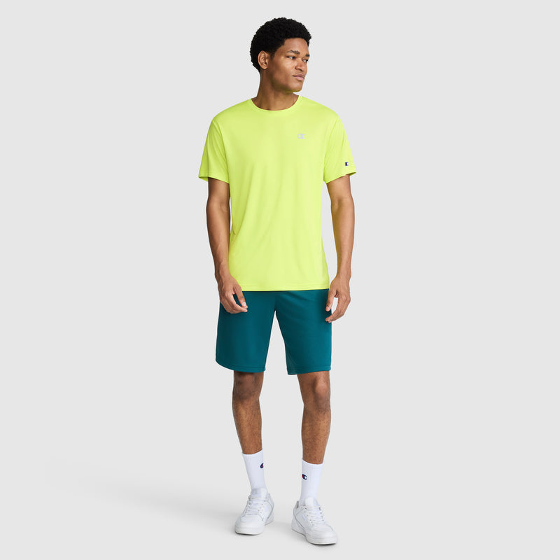 A man stands against a plain light gray background, wearing a bright yellow Champion Sport T-Shirt with a Reflective C Logo, teal shorts, white socks, and white sneakers. He is looking to his left with his arms relaxed by his sides.