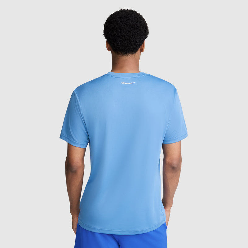 A person stands facing away, wearing a light blue Champion Sport T-Shirt with Reflective C Logo and matching blue shorts. The background is plain light gray.