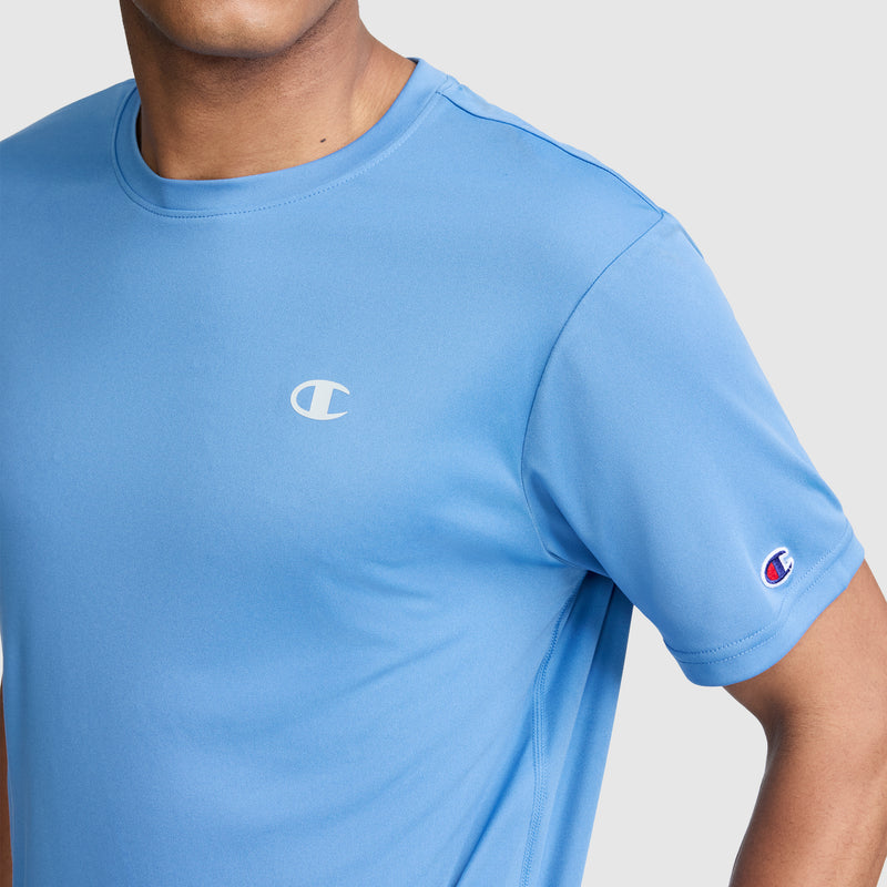 A person wearing a light blue Champion Sport T-Shirt, Reflective C Logo, with the chest logo and sleeve patch visible, their head partially out of frame against a plain background. This men’s Champion athletic shirt delivers both style and comfort.