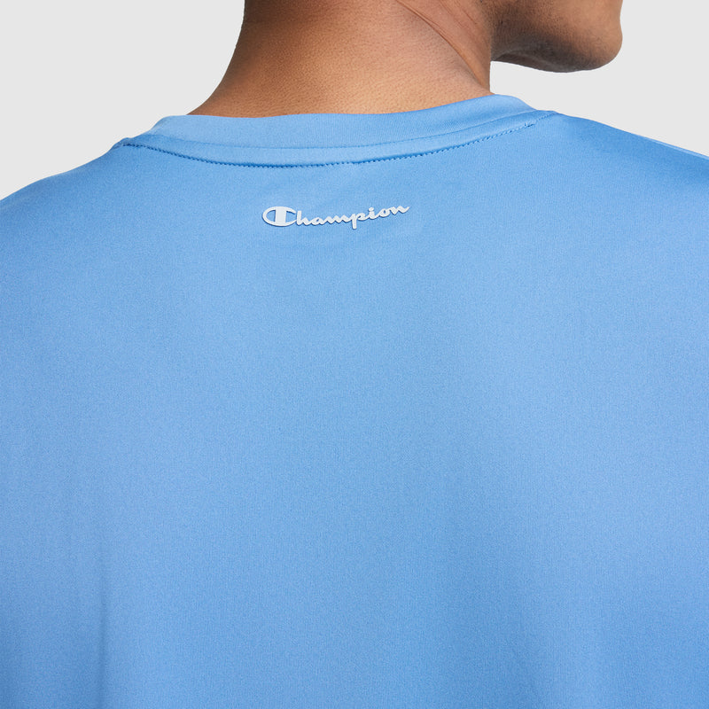 Close-up of a person wearing a light blue Champion Sport T-Shirt, Reflective C Logo, showing the back of the shirt with the white reflective Champion logo near the neckline. The person’s head and neck are partially visible.