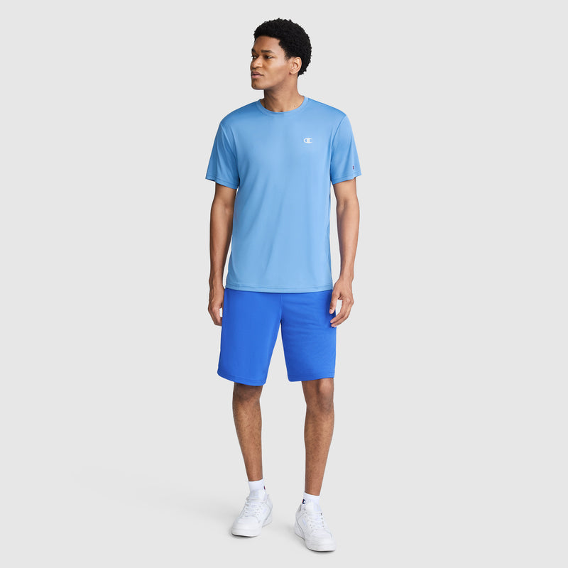 A man stands against a plain light gray background, wearing the Champion Sport T-Shirt with Reflective C Logo in light blue, paired with bright blue shorts and white sneakers.