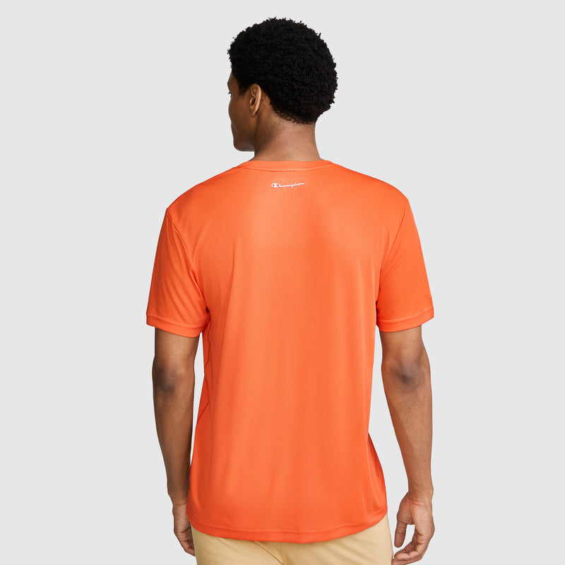 A man is standing with his back to the camera, wearing a Champion Sport T-Shirt, Reflective C Logo in orange and beige pants. The shirt features a small white reflective Champion logo near the neckline. The background is light gray.