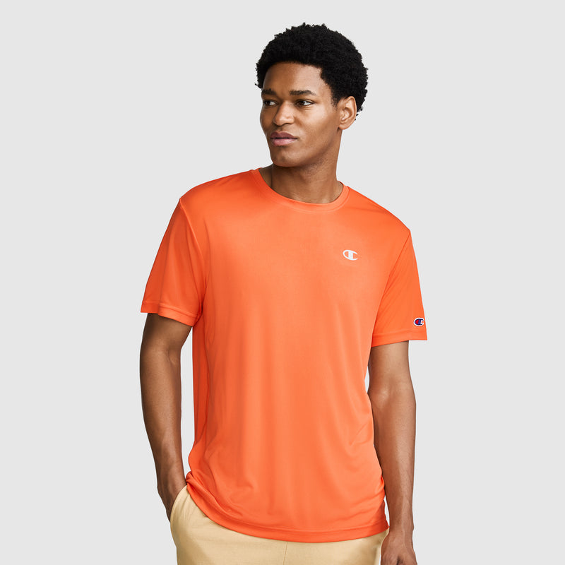A man wearing a Champion Sport T-Shirt with a Reflective C Logo in orange and beige pants stands against a plain gray background, looking slightly to his left with one hand in his pocket.