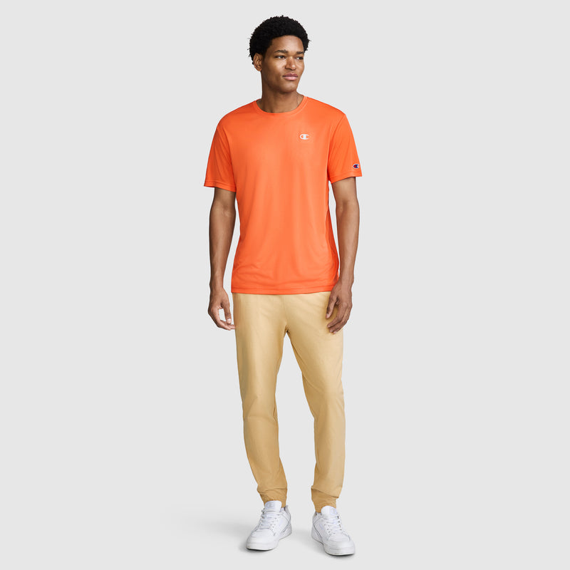 A person stands facing forward, wearing a Champion Sport T-Shirt, Reflective C Logo in orange—featuring moisture-wicking fabric and anti-odor protection—paired with beige pants and white sneakers against a plain light-colored background.