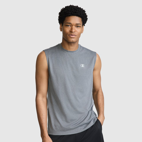 A man wears the Champion Sport Muscle Tank with Reflective C Logo, a sleeveless gray athletic shirt featuring a subtle reflective Champion logo on the chest. Engineered for workouts, its moisture-wicking fabric keeps him comfortable as he stands against a plain light gray background, one hand in his pocket, looking at the camera.