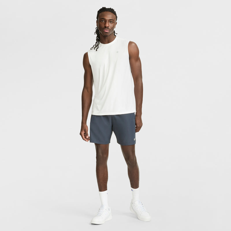 A man with long braided hair stands against a white background wearing a Champion Sport Muscle Tank with Reflective C Logo, dark gray athletic shorts, white crew socks, and white sneakers.