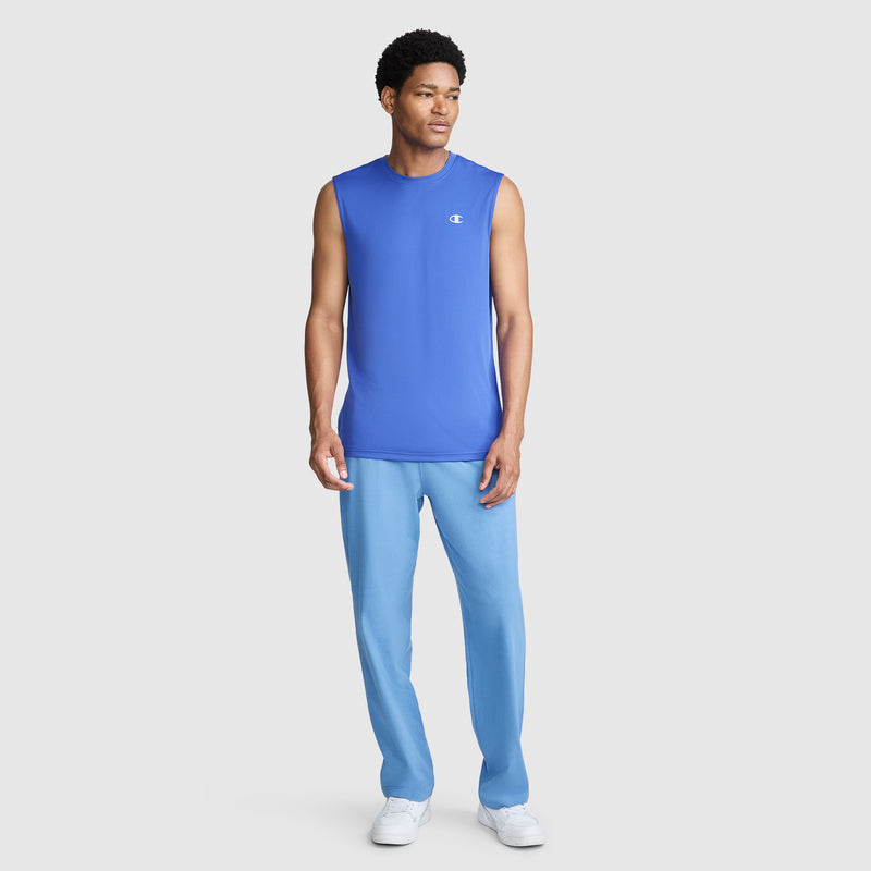A man stands wearing a Champion Sport Muscle Tank with a reflective C logo, paired with light blue pants and white sneakers. The sleeveless workout shirt is made from moisture-wicking fabric as he faces slightly to the side against a plain light background.