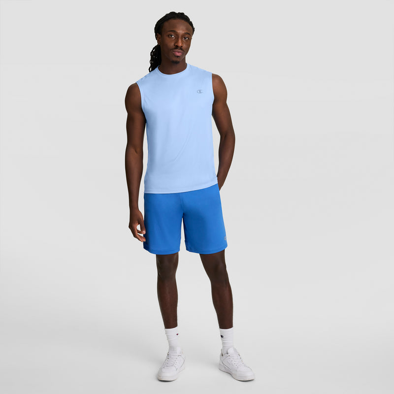 A man with long braided hair and a neutral expression stands against a plain light gray background, wearing the Champion Sport Muscle Tank with Reflective C Logo, blue shorts, white socks, and white sneakers.