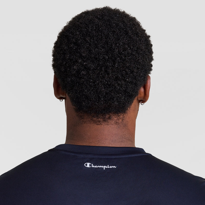 A person with short, curly black hair is shown from behind wearing the Champion Sport Muscle Tank, Reflective C Logo - Big & Tall. The dark workout shirt features a reflective Champion logo on the back. The background is plain and light-colored.