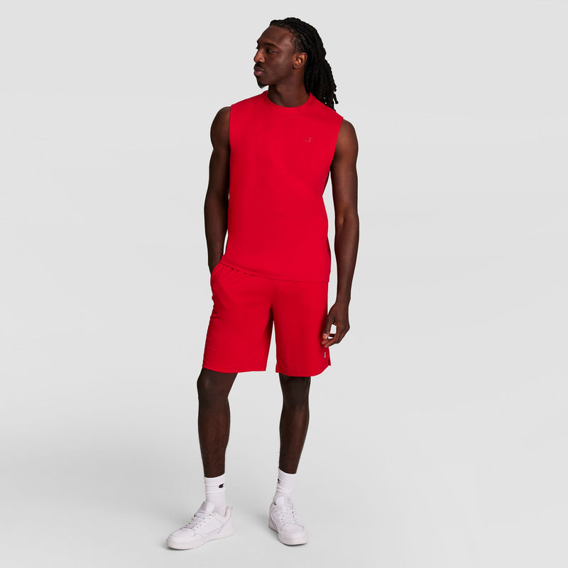 A man in a Champion Muscle Tank, C Logo, and matching red shorts stands with one hand in his pocket, looking to the side. He wears white sneakers and socks. The background is plain light gray.