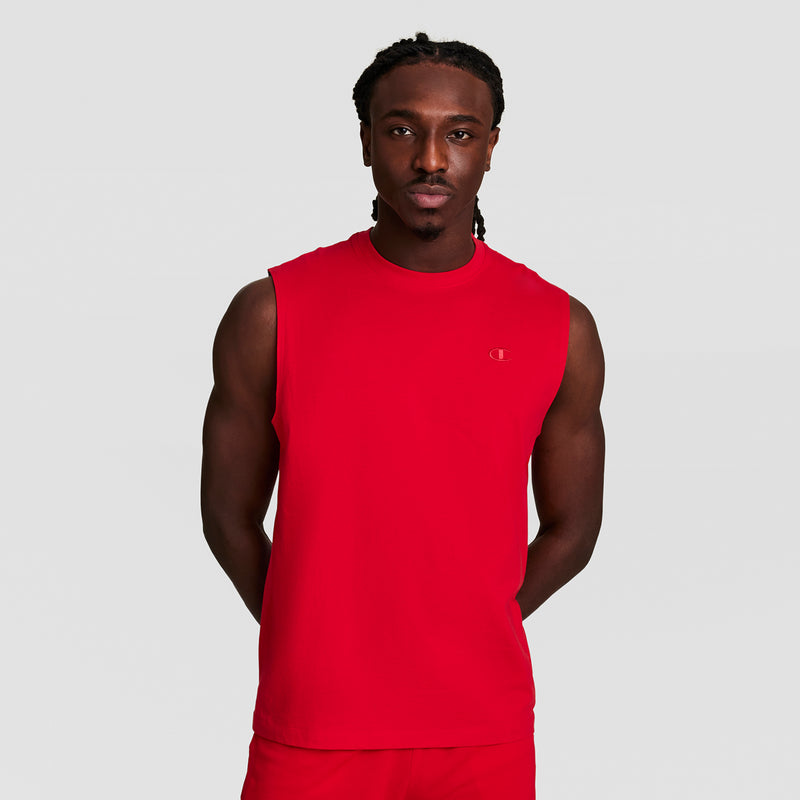 A man with braided hair stands against a plain light gray background, wearing a bright red Champion Muscle Tank featuring the C Logo and matching red shorts, with his hands behind his back.